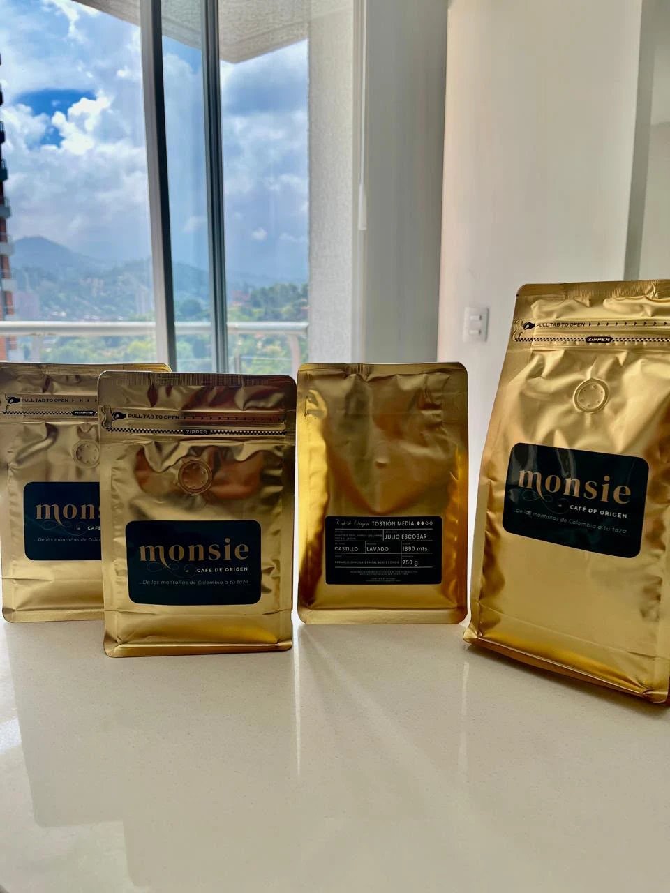 Bolsas doradas de café Monsie frente a una ventana con vista a las montañas de Antioquia